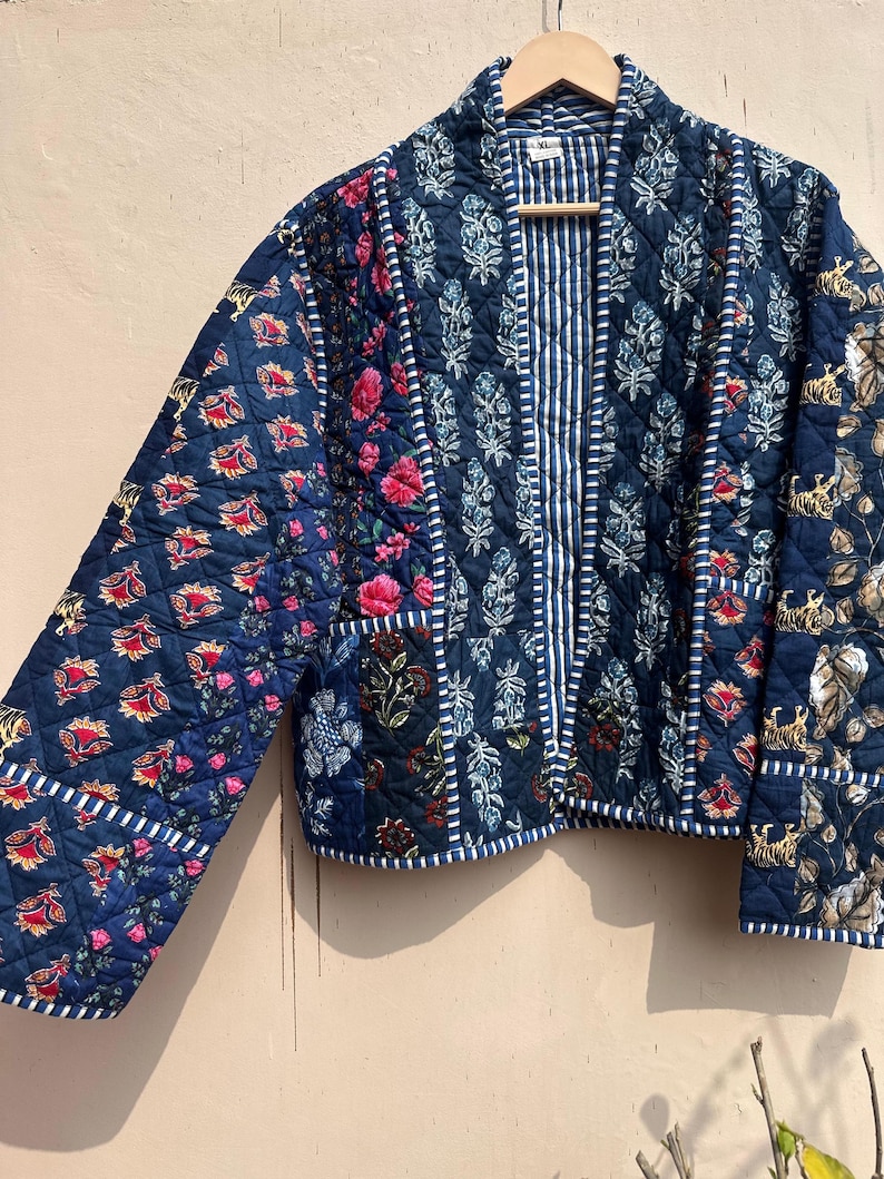 Peut inclure: Veste matelass&eacute;e bleu marine avec patchwork &agrave; imprim&eacute; floral et tigre. La veste pr&eacute;sente un panneau central ray&eacute; et une bordure blanche. Les manches sont longues et la veste est suspendue &agrave; un cintre en bois.