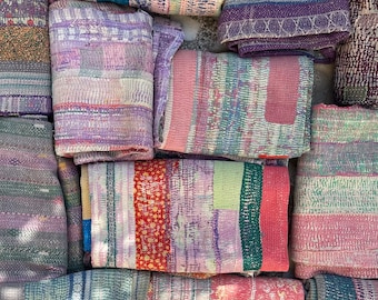 May include: A collection of colorful kantha quilts, folded and stacked on a tiled surface. The quilts are made of patchwork fabric in shades of pink, purple, blue, green, and red. The quilts are hand-stitched and have a vintage, rustic look.