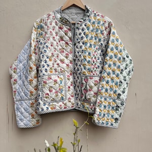 May include: A quilted jacket with a floral pattern in pink, blue, yellow, and green on a white background. The jacket has a black and white striped lining and trim, with a zip closure and two pockets. The jacket is hanging on a wooden hanger.