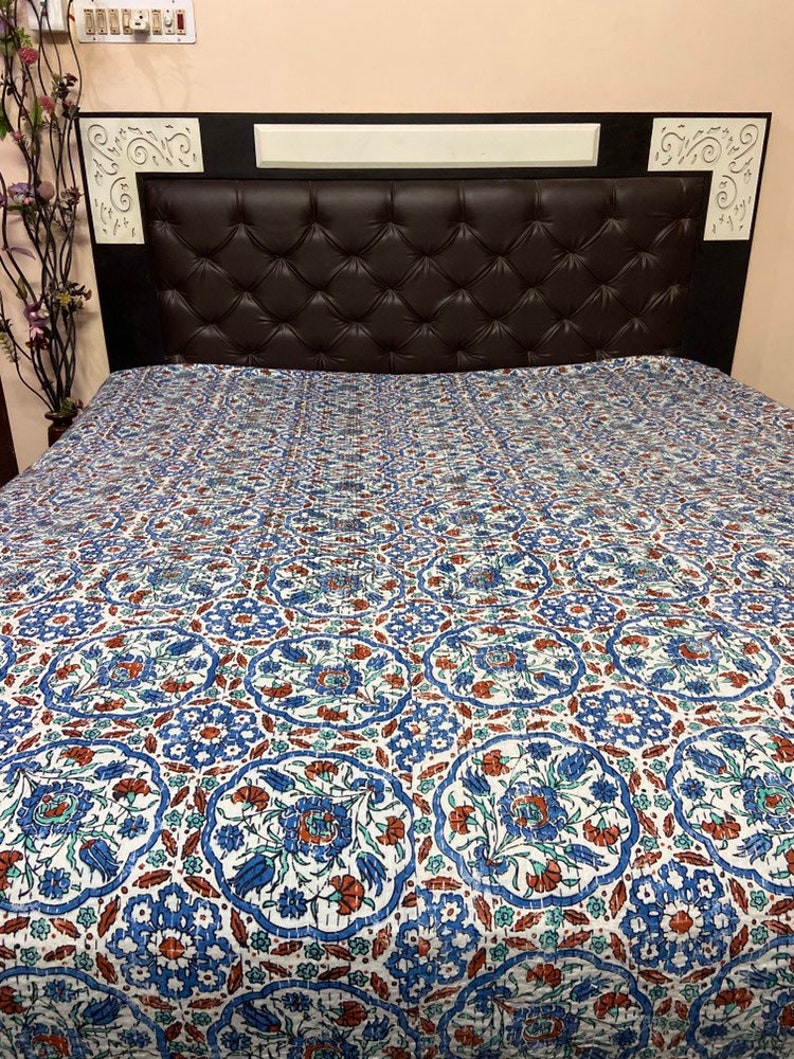 May include: A white cotton quilt with a blue and red floral pattern. The quilt is on a bed with a brown leather headboard.