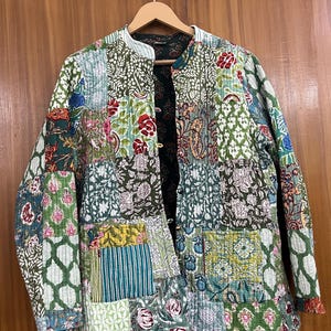 May include: A patchwork jacket with a variety of floral and geometric patterns in shades of green, blue, pink, and yellow. The jacket has a quilted texture, a mandarin collar, and two front pockets. It is hanging on a wooden hanger.
