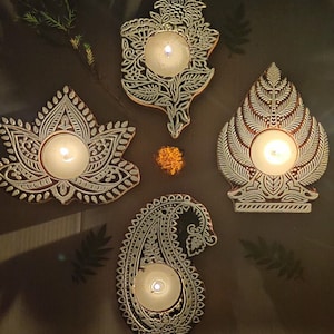 Pode incluir: Quatro porta-velas decorativos com designs intrincados. Cada porta-velas apresenta um padrão diferente, incluindo um paisley, uma flor de lótus, uma árvore e um design floral. Os porta-velas são feitos de um material marrom claro e têm um acabamento branco. Cada porta-velas tem uma pequena cavidade redonda para uma vela de chá.