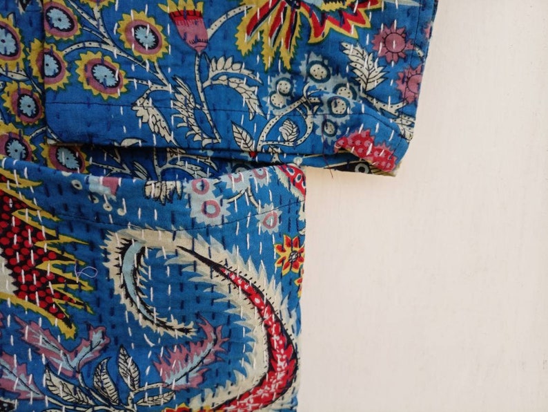 May include: Close-up of a section of a blue quilted fabric with a floral and paisley design. The fabric features a vibrant mix of colors, including red, yellow, and pink, with white stitching. The overall aesthetic is reminiscent of traditional Indian textiles.