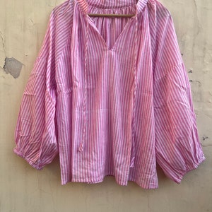 May include: A pink and white striped blouse with a V-neck and long sleeves. The blouse has a tie at the neck and a gathered detail at the shoulders.