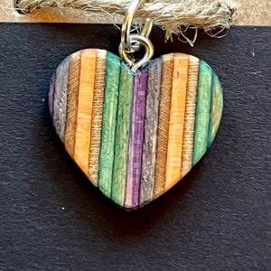 Pode incluir: Um pingente em forma de coração feito de madeira de skate reciclada. O pingente apresenta uma variedade de cores, incluindo verde, roxo, laranja e marrom. O pingente está preso a uma corrente prateada.