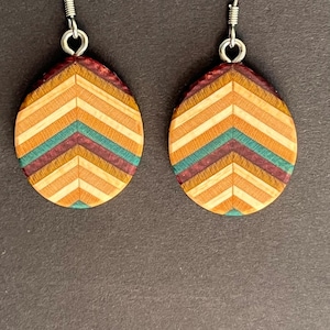 May include: A pair of teardrop-shaped earrings featuring a chevron pattern made from layered wood in shades of light tan, orange, teal, and deep purple. The earrings are attached to silver-toned metal fishhook wires for pierced ears.