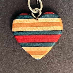Pode incluir: Pingente em forma de coração de madeira com listras horizontais vermelhas, turquesas, laranja e bege. Possui uma argola de metal prateado para pendurar. O fundo é cinza escuro. O pingente tem cerca de 4 cm de largura.