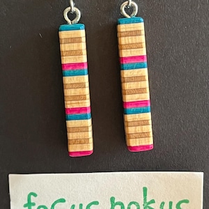 Puede incluir: Un par de pendientes de madera con un patrón de rayas de color turquesa, rosa y madera natural. Los pendientes son de forma rectangular y tienen ganchos plateados.