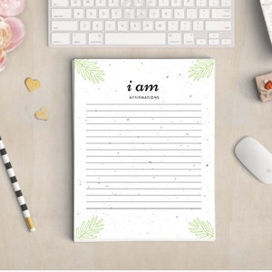 Puede incluir: Un bloc de notas blanco con el texto "i am affirmations" y páginas rayadas. El bloc de notas está decorado con ilustraciones de hojas verdes. También se ven un lápiz a rayas blancas y negras, un teclado de ordenador y una taza de café.
