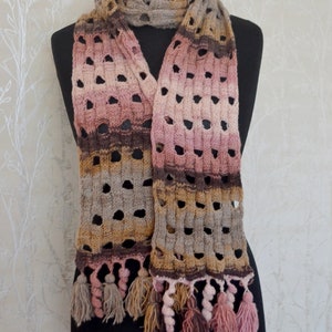 Könnte beinhalten: Ein handgestrickter Schal in Braun-, Rosa- und Beigetönen, präsentiert auf einer schwarzen Schaufensterpuppe. Der Schal hat ein strukturiertes, offenes Webmuster mit rechteckigen Aussparungen und ist mit Quasten an den Enden versehen. Der Schal ist etwa 178 cm lang.