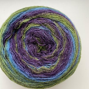 Puede incluir: Una madeja de hilo multicolor, con tonos verdes, azules y morados. El hilo está bien enrollado, con un efecto degradado visible. Adecuado para proyectos de tejido o ganchillo, el hilo ofrece una mezcla de colores.