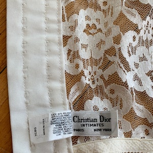 Może przedstawiać: Biała koronkowa bielizna z białą metką z napisem "Christian Dior Intimates Paris New York" i białą metką z instrukcjami pielęgnacyjnymi. Metka zawiera napis "Koronka: 60% poliester 40% nylon Panel Importowany poliester Elastyczny: 79% nylon Spandex Pranie w pralce w ciepłej wodzie z podobnymi kolorami. Tylko wybielacz bez chloru, jeśli to konieczne. Suszenie w suszarce bębnowej w niskiej temperaturze. RN 59993"