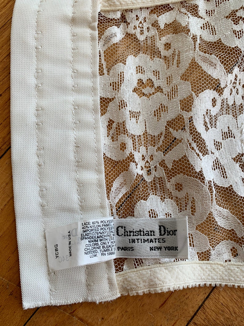 Może przedstawiać: Biała koronkowa bielizna z białą metką z napisem "Christian Dior Intimates Paris New York" i białą metką z instrukcjami pielęgnacyjnymi. Metka zawiera napis "Koronka: 60% poliester 40% nylon Panel Importowany poliester Elastyczny: 79% nylon Spandex Pranie w pralce w ciepłej wodzie z podobnymi kolorami. Tylko wybielacz bez chloru, jeśli to konieczne. Suszenie w suszarce bębnowej w niskiej temperaturze. RN 59993"