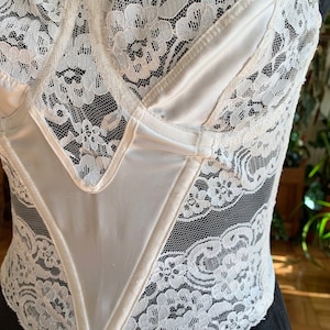 Może przedstawiać: Biały gorset z satyny i koronki z dekoltem w kształcie serca. Gorset ma sznurowaną konstrukcję i jest ozdobiony delikatną koronką.