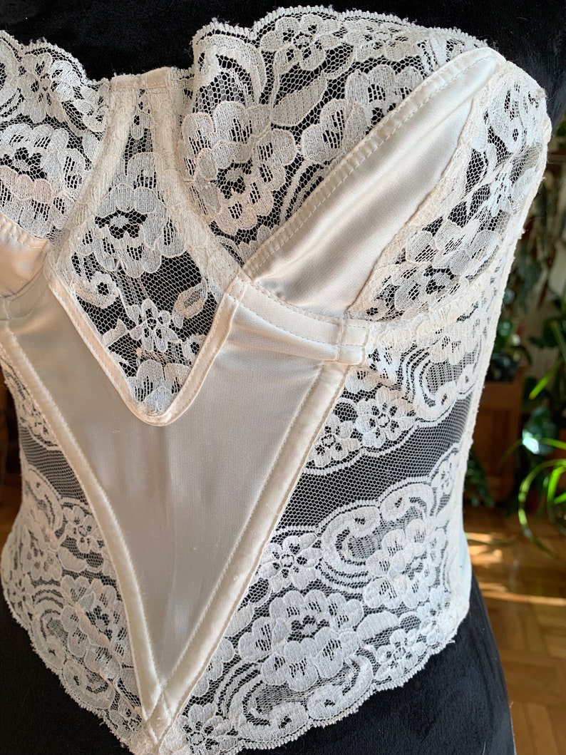 Może przedstawiać: Biały gorset z satyny i koronki z dekoltem w kształcie serca. Gorset ma sznurowaną konstrukcję i jest ozdobiony delikatną koronką.