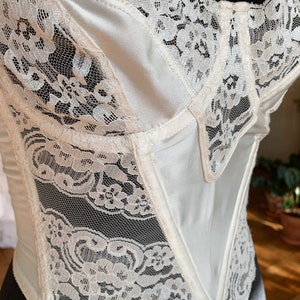 Może przedstawiać: Biały gorset z satyny i koronki z ostrym przodem i falbanką. Gorset wykonany jest z delikatnej koronki z kwiatowym wzorem.