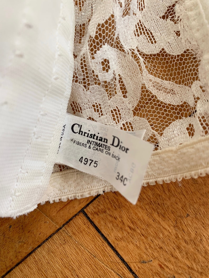 Może przedstawiać: Biała koronka etykieta biustonosza z tekstem "Christian Dior Intimates" i rozmiarem "34C". Etykieta zawiera r&oacute;wnież numer "4975" i tekst "Fibers & Care on Back".