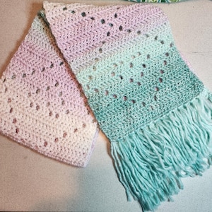 May include: A crocheted scarf with a gradient of pink and green colors. The scarf has a fringe on the bottom and is made with a delicate stitch pattern.