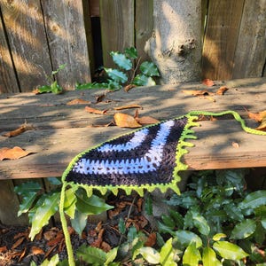 May include: A handmade crochet bandana in black, white, and light blue stripes, outlined with bright green. The bandana is triangular with a long green tie, resting on a wooden bench with a natural background.
