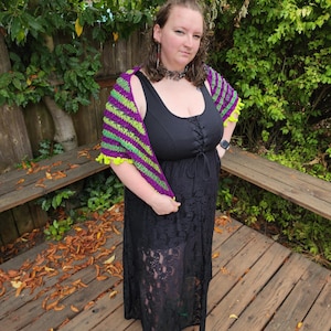 May include: A handmade crochet shawl with alternating purple and green stripes. The shawl has a ruffled neon yellow trim. The person in the image is wearing a black dress and green shoes. The background includes a wooden deck and green foliage.