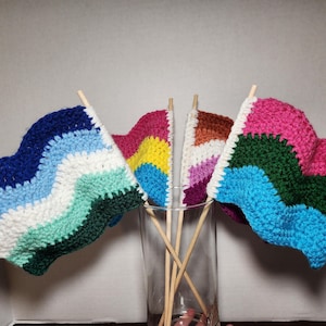 May include: Four handmade crochet flags in a clear glass. The flags are attached to wooden dowels. The flags feature various color schemes, including blue, white, green, pink, yellow, and brown. The flags are likely decorative items.
