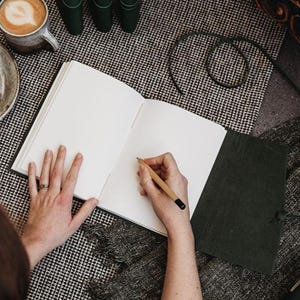 May include: An open, blank journal with a dark green leather cover and a pencil. The journal is on a black and white checkered surface, with a cup of coffee and a plate in the background. The pencil is being held by a person.