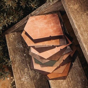 May include: A stack of leather pieces in various shades of brown, tan, and olive green. The pieces are hexagonal with a notched design. The leather has a textured appearance, and the pieces are arranged on a weathered wooden surface.