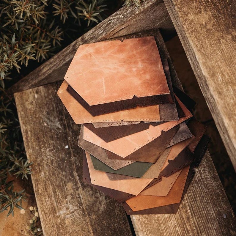 May include: A stack of leather pieces in various shades of brown, tan, and olive green. The pieces are hexagonal with a notched design. The leather has a textured appearance, and the pieces are arranged on a weathered wooden surface.