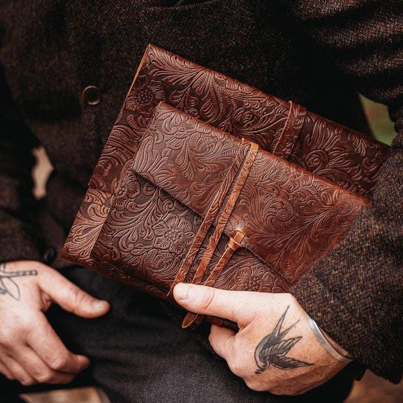 May include: Two brown leather journals with embossed floral patterns. The larger journal is open, revealing the interior. The smaller journal is closed with a leather strap. The journals are held together.