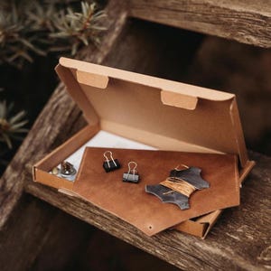 May include: A brown leather wallet with black binder clips and a dark gray leather piece with twine, inside a cardboard box. The box is open, revealing the contents on a wooden surface.