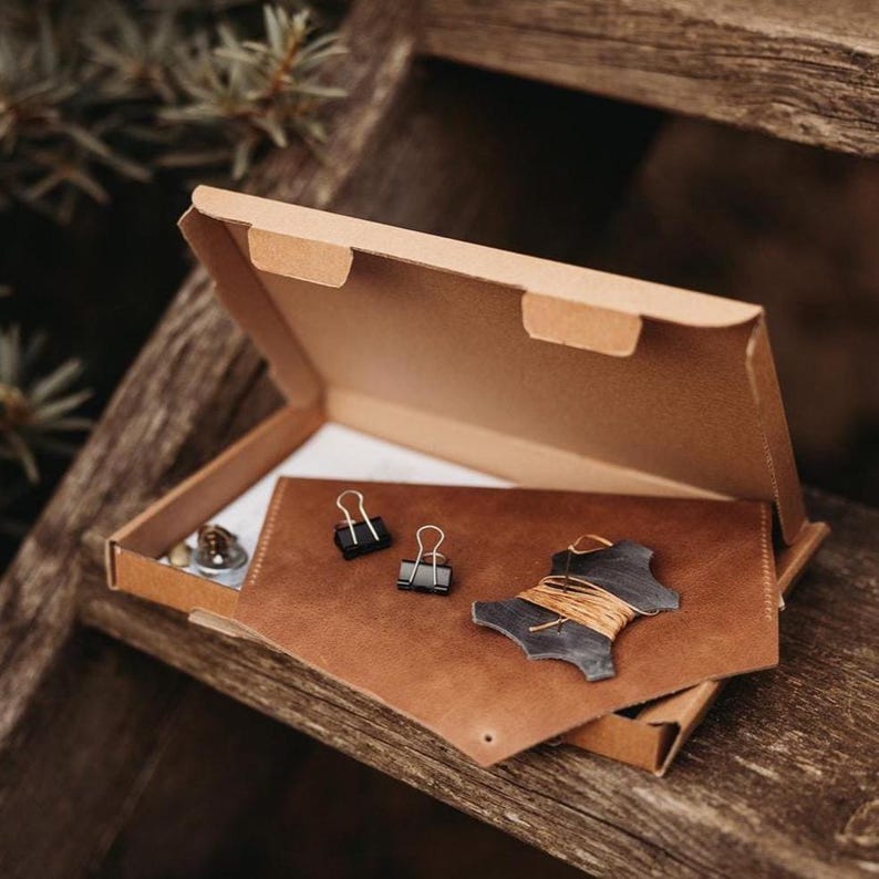 May include: A brown leather wallet with black binder clips and a dark gray leather piece with twine, inside a cardboard box. The box is open, revealing the contents on a wooden surface.