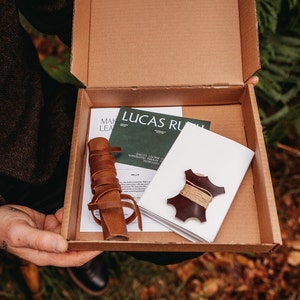May include: A brown cardboard box with the lid open, revealing a leather craft kit. The kit includes a piece of leather, a leather strap, and instructions. The box is labelled "Lucas Ruth" and "Make Leather"
