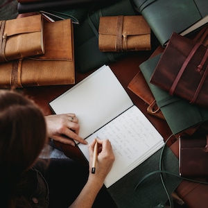 Puede incluir: Una persona escribiendo en un diario en blanco con un lápiz de madera. El diario está rodeado de varios diarios encuadernados en cuero de diferentes colores y tamaños.