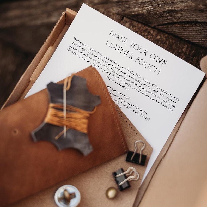 May include: A leather pouch making kit in a cardboard box. The kit includes a brown leather pouch, a piece of leather with thread, paper instructions, and metal fasteners. The text on the paper reads "MAKE YOUR OWN LEATHER POUCH".