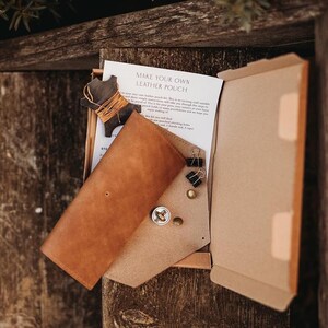 May include: A leather pouch kit with a brown leather pouch, a tan leather piece, and a paper with the text "MAKE YOUR OWN LEATHER POUCH". The kit includes a wooden shape wrapped with orange thread, metal fasteners, and binder clips.