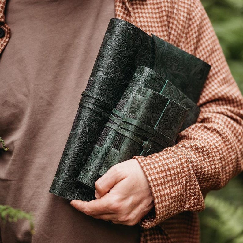 May include: Dark green leather journals with embossed floral patterns. The journals are bound with leather straps. The person is wearing a brown and tan houndstooth jacket and a brown shirt.