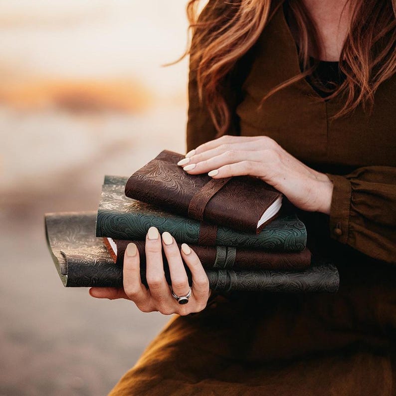 May include: A stack of leather-bound journals in varying shades of brown and green. The top journal is a dark brown with a strap closure. The covers feature embossed floral patterns. The journals are held in a person's hands.