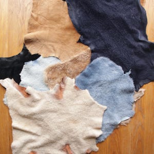 May include: A selection of irregularly shaped, tanned leather hides in shades of tan, black, light blue, and cream. The hides are displayed on a wooden surface, highlighting their natural textures and colour variations.