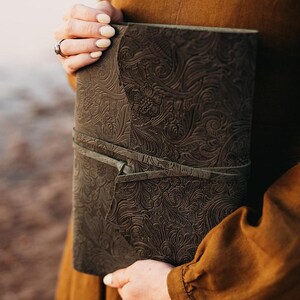 May include: A dark brown leather journal with an embossed floral pattern. The journal is held closed by a matching leather strap. The cover is intricately detailed with a repeating design of flowers and leaves.
