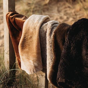 May include: Several faux fur throws in shades of brown and cream are draped over a wooden fence. The textures vary from smooth to shaggy. The throws are displayed outdoors, with a range of colours from light tan to dark brown.