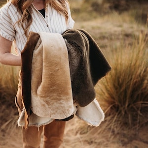 May include: A collection of three shearling blankets in brown, beige, and olive green. The blankets have a soft, textured appearance. A person is holding the blankets, wearing a striped shirt and brown trousers. The background shows dry grass.