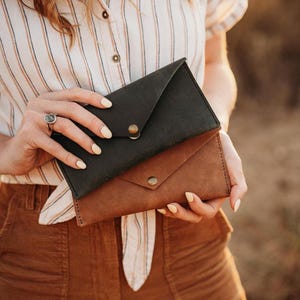 May include: Two leather envelope-style wallets, one dark green and one brown, are held. Each wallet has a flap closure secured by a brass-colored snap. The wallets are made of a textured leather material.