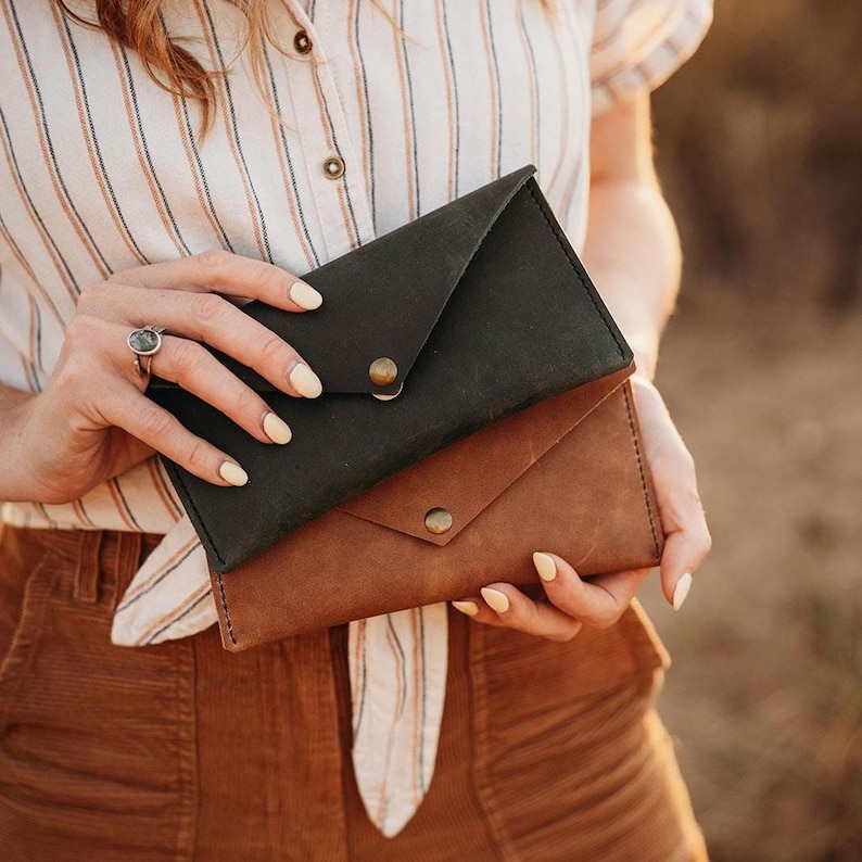 May include: Two leather envelope-style wallets, one dark green and one brown, are held. Each wallet has a flap closure secured by a brass-colored snap. The wallets are made of a textured leather material.