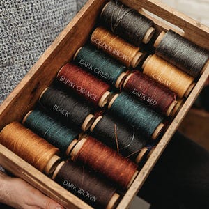 May include: A wooden box filled with spools of thread in various colors, including golden brown, dark green, burnt orange, black, dark red, and dark brown. Each spool is labeled with its color name. The thread spools are arranged neatly.