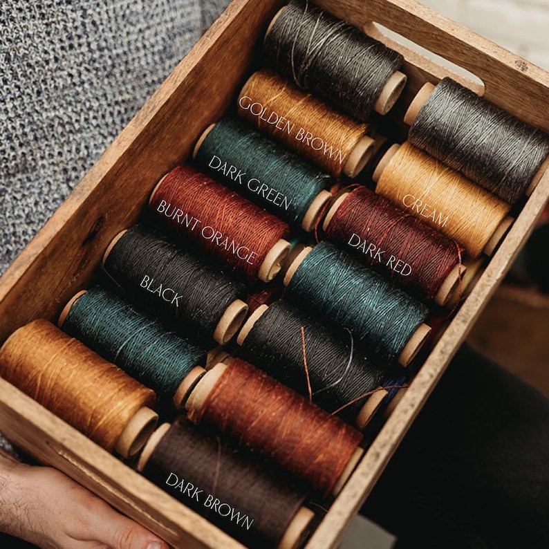 May include: A wooden box filled with spools of thread in various colors, including golden brown, dark green, burnt orange, black, dark red, and dark brown. Each spool is labeled with its color name. The thread spools are arranged neatly.