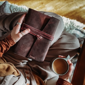 May include: A person is sitting in a chair holding a brown leather journal with the word "Dream" embossed on the cover. They are also holding a cup of coffee.
