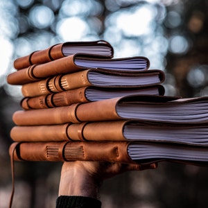 Op de afbeelding: Een stapel van zes lederen gebonden notitieboeken met crèmekleurige pagina's. De notitieboeken hebben een warme bruine kleur en een eenvoudig, klassiek ontwerp. De pagina's worden bij elkaar gehouden met stiksels langs de rug. De notitieboeken worden in een hand gehouden.