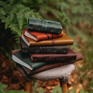 Peut inclure: Une pile de carnets en cuir de différentes couleurs, dont vert, marron et orange. Certains carnets présentent des motifs floraux en relief et des lanières en cuir. Les carnets sont empilés sur un petit tabouret rustique, avec un fond vert flou.
