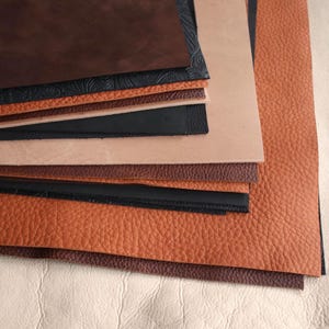 May include: A selection of leather samples in a range of browns, tans, and black. The textures vary from smooth to textured, demonstrating the different finishes. The leather pieces are arranged on a light-coloured surface.