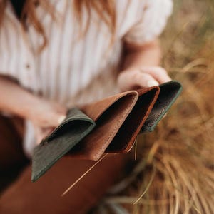 May include: A selection of leather wallets in various colors, including dark green, brown, and reddish-brown. The wallets are held in a person's hands, showcasing their different textures and hues. The wallets appear to be new and unused.